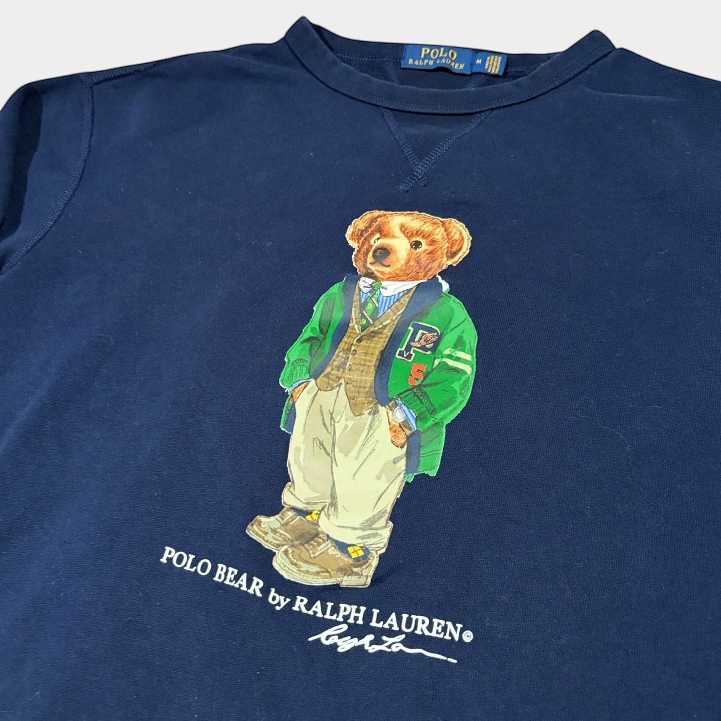 THE YRDSALE navy crewneck sweatshirt with a graphic of the Polo Bear by Ralph Lauren wearing a green varsity jacket and tan trousers.